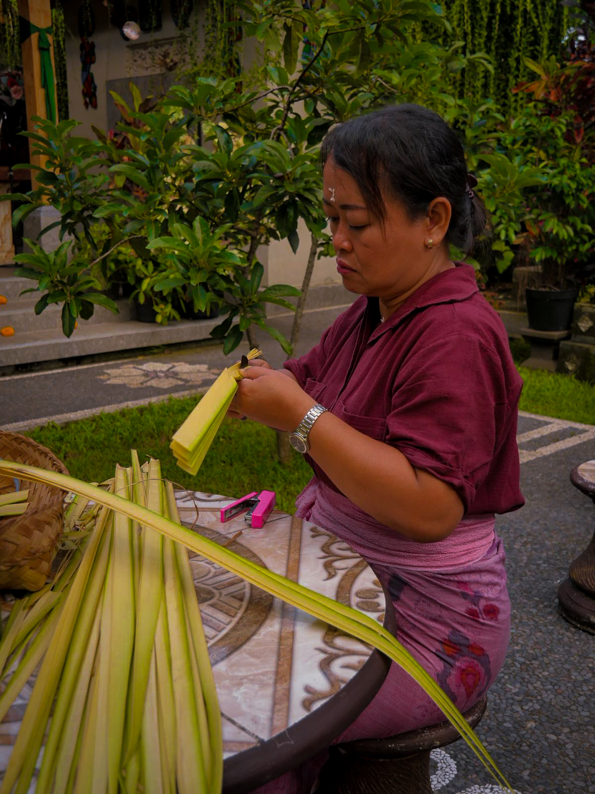 donna balinese seduta in un giardino mentre costruisce dei canang sari tagliando foglie di palma